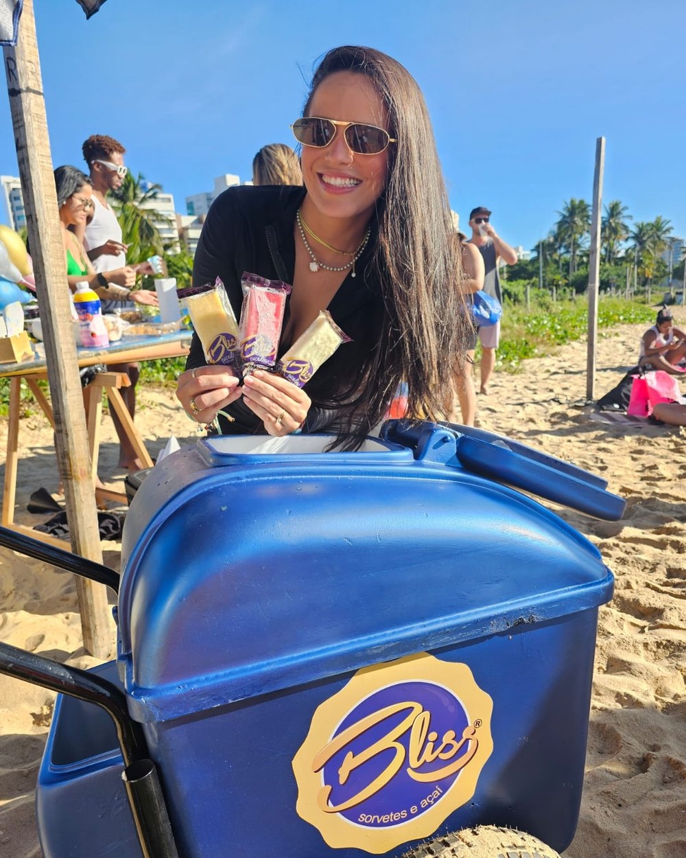 Atendente da Bliss segurando três picolés ao lado do carrinho azul da marca, na areia da praia